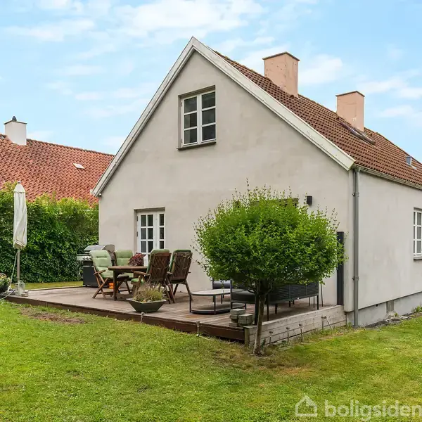 Et hvidt hus med rødt tag står i en grøn have. På terrassen er der havemøbler og en parasol. Hække omkranser græsplænen.