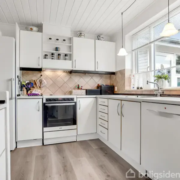 Et hvidt køkken med ovn og skabsplads placeret i et lyst rum med trægulv. Vindue og hvide loftlamper giver naturligt lys. Hylder med krus og planter dekorerer rummet.