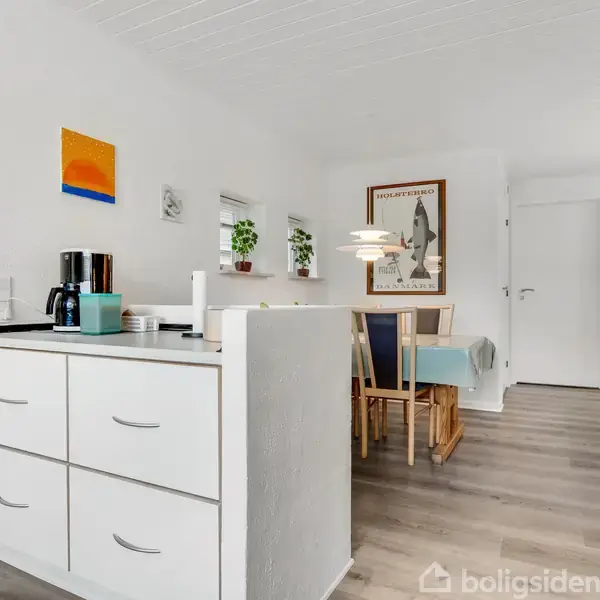 Køkkenbord med kaffemaskine og kopper, placeret i et lyst køkken. I baggrunden ses et spisebord med stole og vægdekorationer i en moderne, minimalistisk indretning.