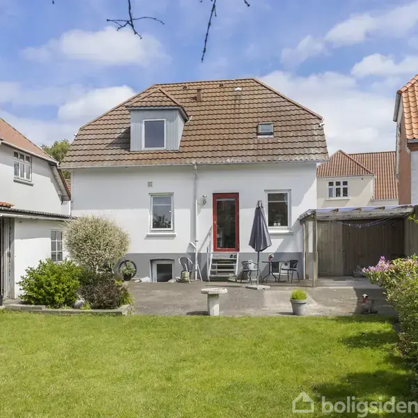 Hvidt hus med rødt tag står i en have med græsplæne og fliseterrasse. På terrassen er havemøbler og parasol. Omkring huset ses andre lignende bygninger under en blå himmel.