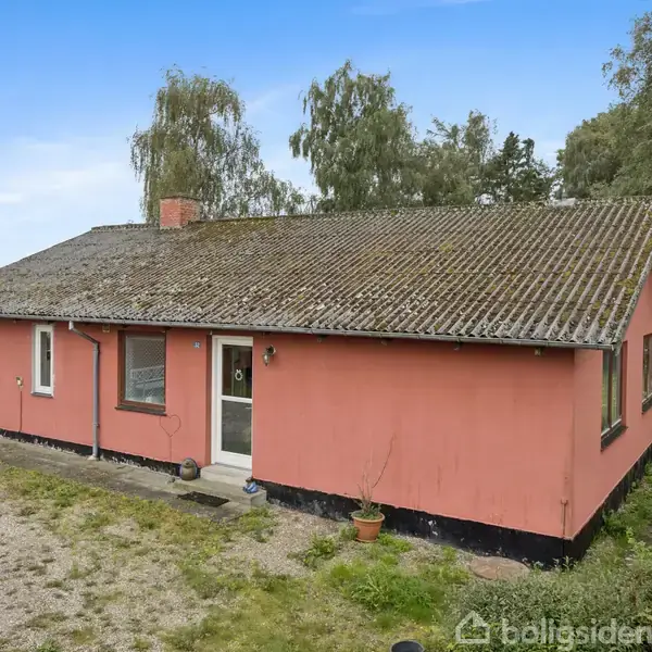 Rødt hus med mosdækket tag og tre vinduer. Det står i grønne omgivelser med træer og græs, og himlen er klar blå. Der er en grussti foran huset.