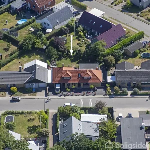 Et boligområde ses oppefra. En pil peger mod et rødt taghus i midten. Omkringliggende huse og haver er synlige. Vej med biler og cyklister foran husene.