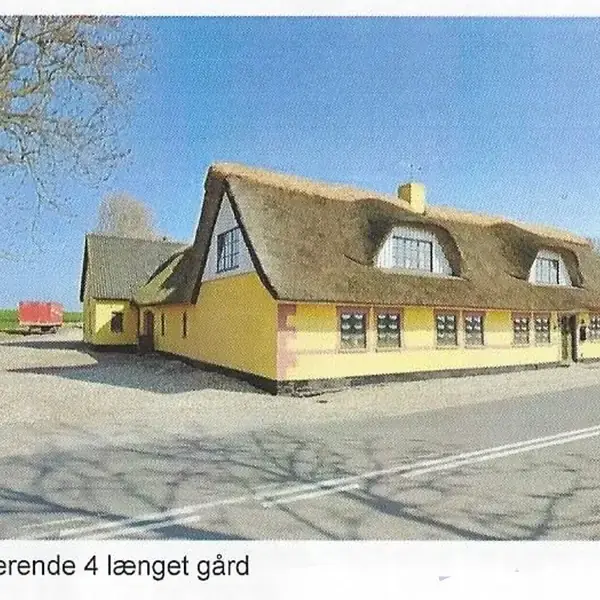 En gul, stråtækt gård står langs en vej, omkranset af åbne marker og træer. Skylinen er blå. Tekst: "Charmerende 4 længet gård".