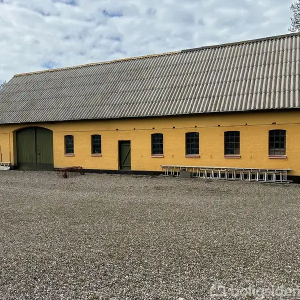 En gul bygning med grønne døre står på en grusplads. Flere stiger er placeret langs væggen. Træer og himmel ses i baggrunden.