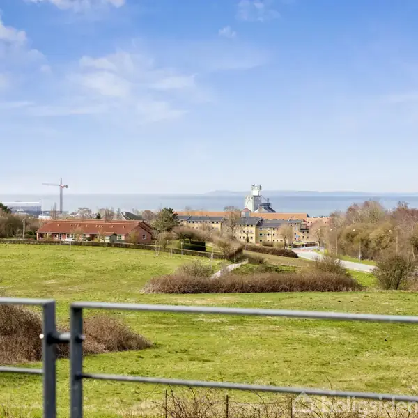 Grøn mark strækker sig mod en fjern bygning med et hvidt tårn. I baggrunden ses blå himmel og vand. Træer og buske omkranser landskabet.