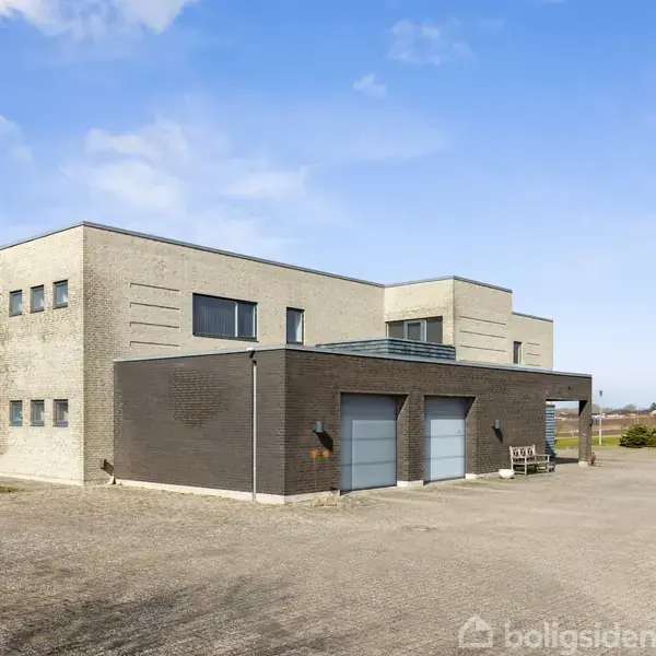 En moderne, kubistisk bygning med fladt tag og store vinduer står på en grusbelagt grund omgivet af grønne marker under en blå himmel med lette skyer.