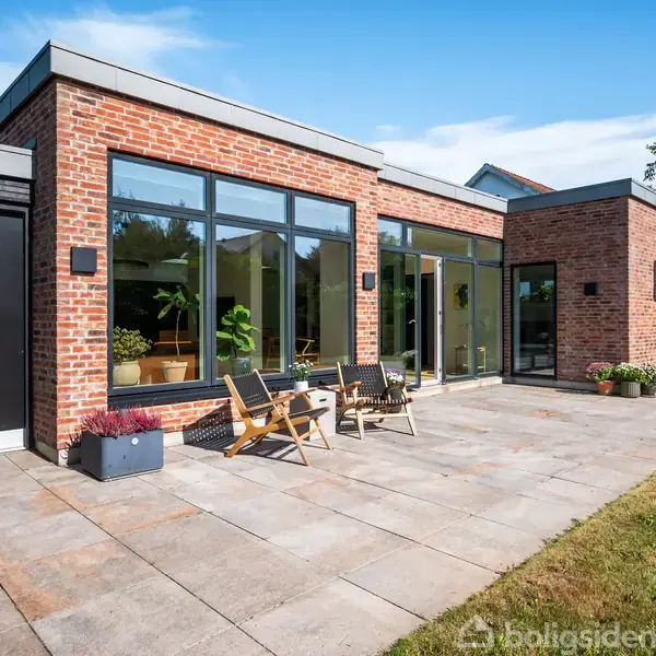 Murstenshus med store vinduer og terrasse, hvor to stole er placeret. Haven omkranser terrassen med grønne planter og græsplæne. En lys og solrig dag skaber en indbydende atmosfære.