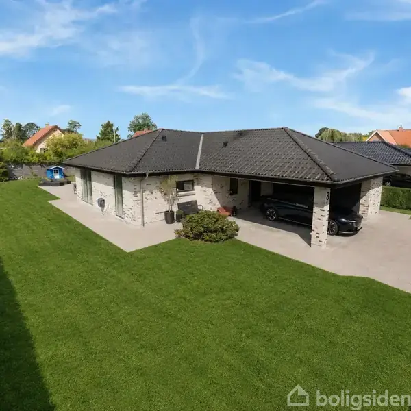 En moderne villa med fladt sort tag står i en velplejet have med grøn græsplæne. En bil er parkeret i en integreret carport. Omkringliggende hække og nabohuse ses.