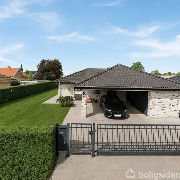 En moderne villa med gråt tag og muret facade står bag en sort bil parkeret i en dobbeltgarage. Huset er omgivet af en velplejet græsplæne og hæk; nummer 8 ses tydeligt.