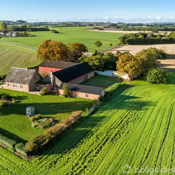 En firlænget gård med stald og hovedbygning omgivet af grønne marker og træer. Beliggende i et landligt område med spredte små landsbyer i baggrunden.
