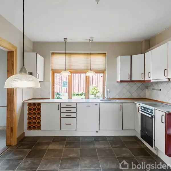 Køkken med hvide skabe og træhylder, der omslutter en ovn. To lamper hænger over bordpladen. Store vinduer med persienner giver lysindfald. Flisegulv og åbning til tilstødende rum.