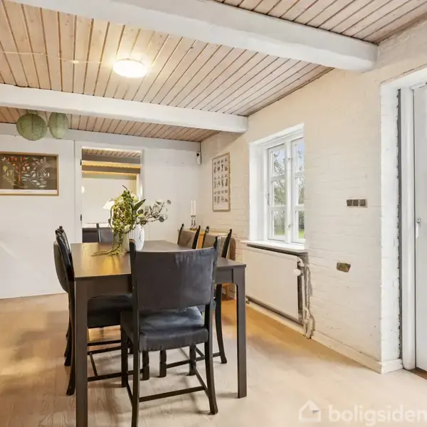 Et spisebord med sorte stole står i et lyst køkkenalrum. Bordet er dekoreret med en vase med blomster, og store vinduer giver udsigt til en grøn have udenfor.