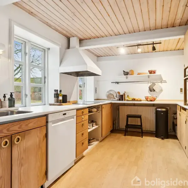 Et køkken har træskabe og hvid bordplade med en indbygget vask. Emhætte over kogeplade. Reol med køkkenudstyr. Vindue giver naturligt lys. Trægulv og træloft fuldender rummet.