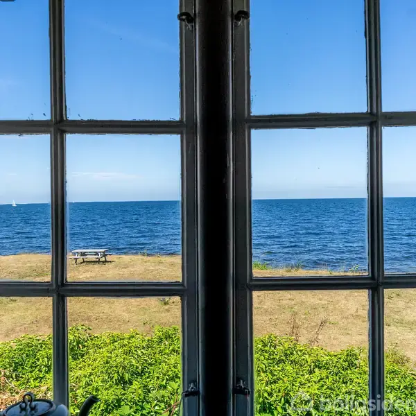 Et vindue åbner op til en udsigt over havet, med en græsplæne og en bænk i forgrunden, omgivet af grønne buske under en klar blå himmel.