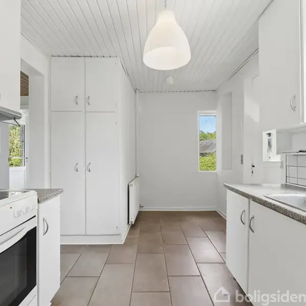 Hvidt køkken med ovn og vask på hver sin side, overskabe og hvidt loft. Store fliser på gulvet, hængende lampe fra loftet, vindue i baggrunden med udsigt til grønne træer.