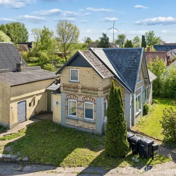 Hus med gul mursten står i en have med græs og buske. Bygningen har skråtag og vinduer. Det ligger i et beboelseskvarter med flere huse og træer omkring.