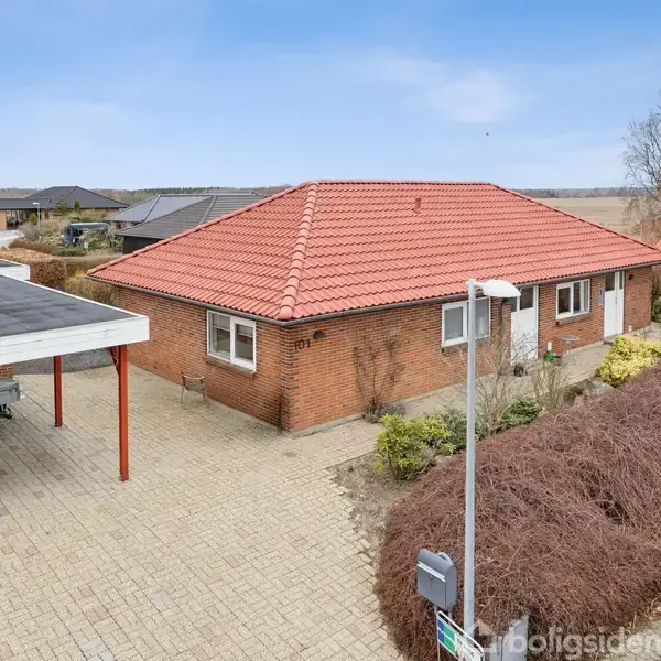 Et rødt murstenshus med rødt tegltag står stille ved siden af en carport. Omgivet af flisebelagt indkørsel og buskads i et boligområde.
