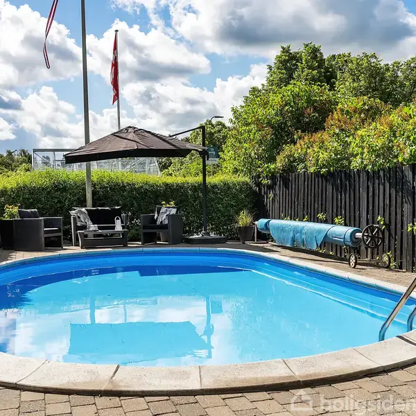 Swimmingpool står stille i et flisebelagt gårdmiljø. Omkring er der grøn buskads og en sort træhegn. Flagstang med flag og havemøbler med parasol står ved siden af.