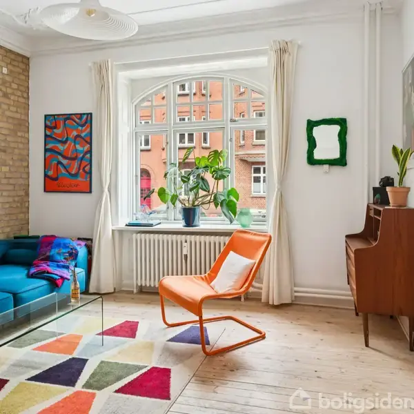 En orange stol står foran et stort vindue med planter, omgivet af en blå sofa og et klaver. Værelset har trægulv og farvestrålende kunst på væggen.