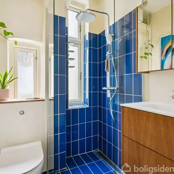 Et moderne badeværelse med blå fliser i brusekabinen, en hvid vask på et træskab, og et toilet. Der hænger en farverig plakat og en plante over vinduet.