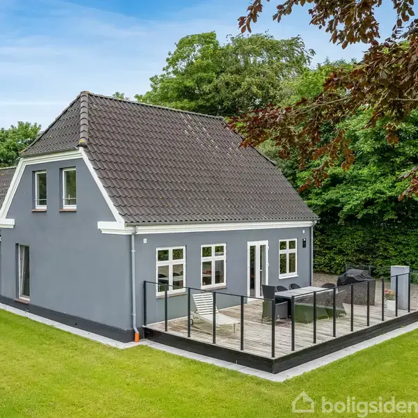 Et gråt hus med hvide vinduesrammer står på en grøn græsplæne. Terrassen er afskærmet med glasgelænder. Huset omkranses af træer og buske, hvilket skaber en privat atmosfære.