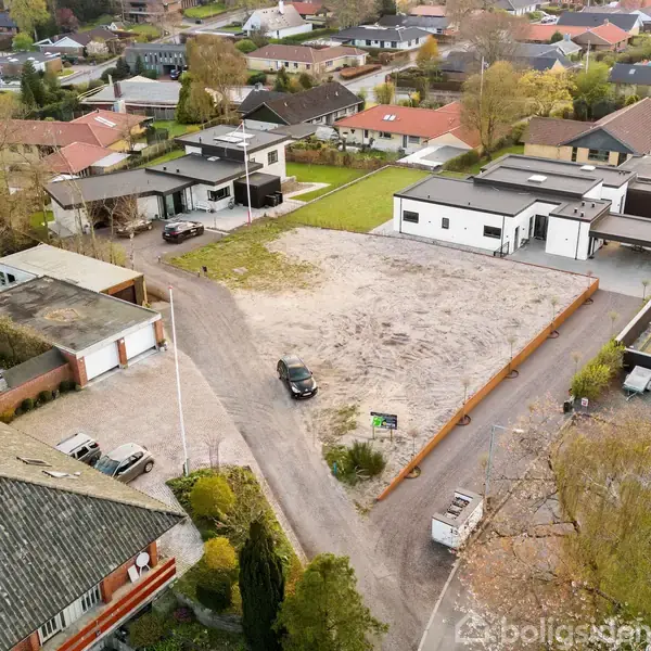 En åben byggegrund ligger mellem moderne huse i et boligområde. En bil holder parkeret på jorden. Grunden er omkranset af veje og haver, mens grønne træer ses omkring.
