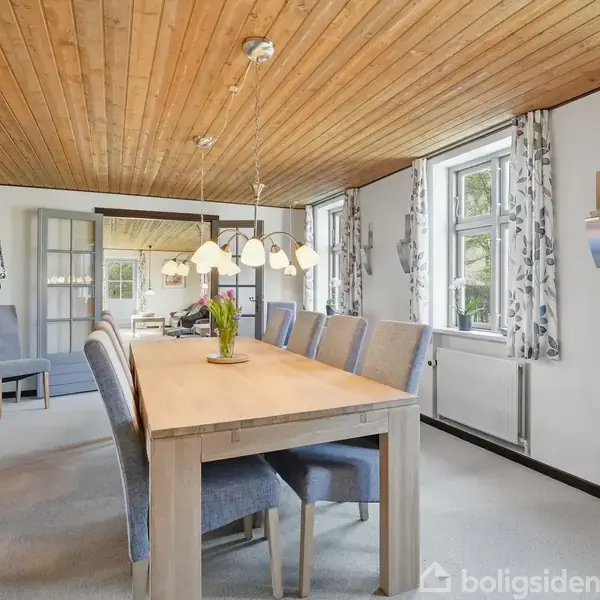 Langt spisebord med blå stole står i en lys spisestue. Træloft og store vinduer prydet med mønstrede gardiner. En træskænk står i baggrunden. Bordet har en vase med blomster.