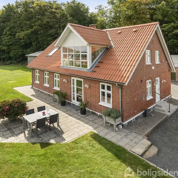 Et rødt murstenshus med rødt tag står i sollys, omgivet af en stor have. Ved siden af huset er en terrasse med et havesæt på fliser.