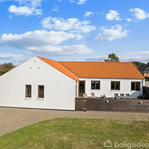 Hvidt hus med rødt tegltag står stille omgivet af en gårdhave med fliser og grøn græsplæne. Ved siden af er der et mørkt skur under en blå himmel med lette skyer.