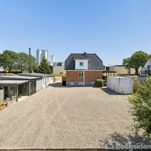 Et lille hus med sort tag står på en grusbelagt gårdsplads. Omkring huset er der flere skure og en garage, mens grønne træer og bygninger ses i baggrunden.