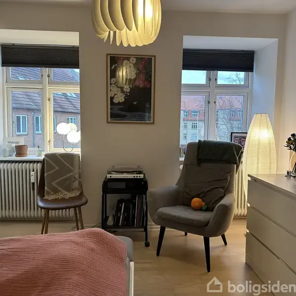 Stol med pude er placeret i en lys stue ved siden af et højt, tændt gulvlampe. To vinduer med udsigt til bygninger udgør baggrunden, mens et tæppe hænger på en stol.