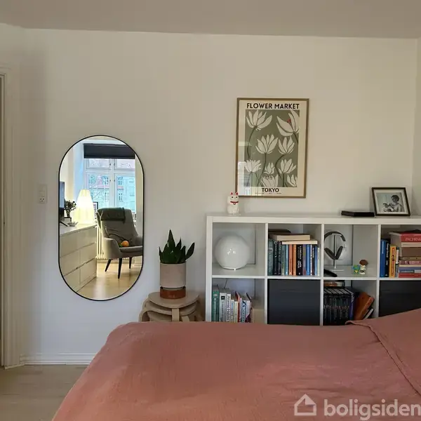 En seng dækket med et lyserødt sengetæppe står i et soveværelse med en hvid væg. Over sengen hænger et spejl. Reolen ved siden af har bøger og dekorationer. På væggen hænger en plakat med teksten "FLOWER MARKET TOKYO".