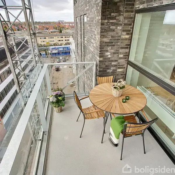 Balkon med et rundt træbord omgiven af fire stole. Ved siden af står en plante. Udsigt over bymiljø med fleretagers bygninger i baggrunden.