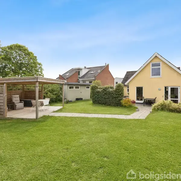 Et gult hus med en enkelt etage og loftsvindue. En grøn have omgiver huset. Til venstre er en overdækket terrasse med møbler. Blå himmel ses i baggrunden.