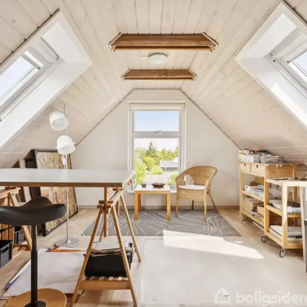 Et lyst, isoleret rum med skrålofter og to ovenlysvinduer. Et tegnebord står midt i rummet med en stol, mens en lænestol og hylder ses i baggrunden.