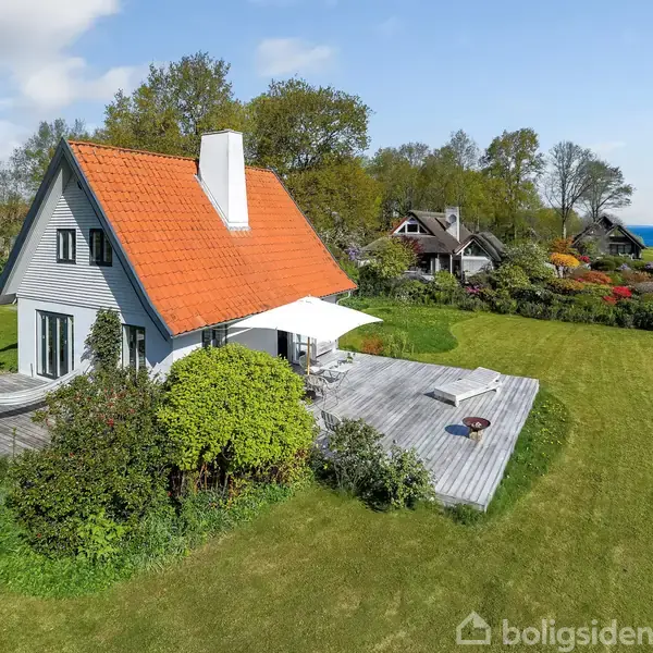 Et hvidt hus med rødt tag ligger stille omgivet af en stor grøn have. Det står på en træterrasse og omgives af frodige buske. Havet ses i baggrunden.