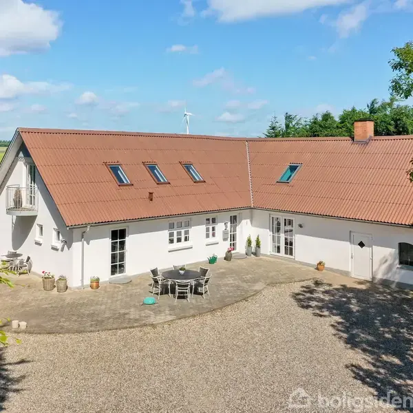 Et hvidt, L-formet hus med rødt tag står stille omgivet af grusbelagt gårdsplads, grønne marker, træer og en vindmølle i baggrunden under en blå himmel med skyer.