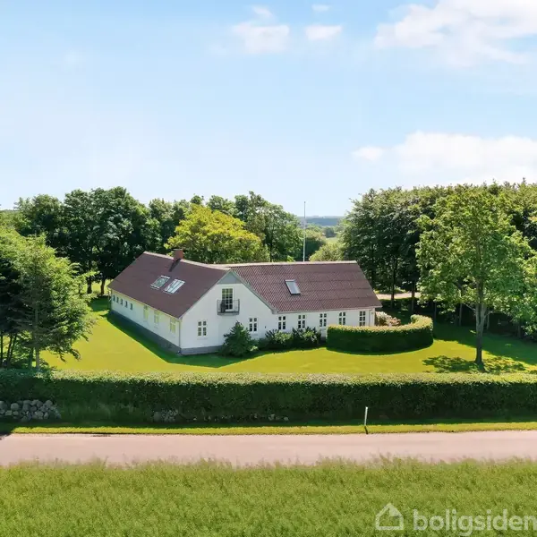 Hvidt hus med rødt tag står stille omgivet af en velplejet have og høje træer. En vej og grøn eng er i forgrunden under en blå himmel med få skyer.