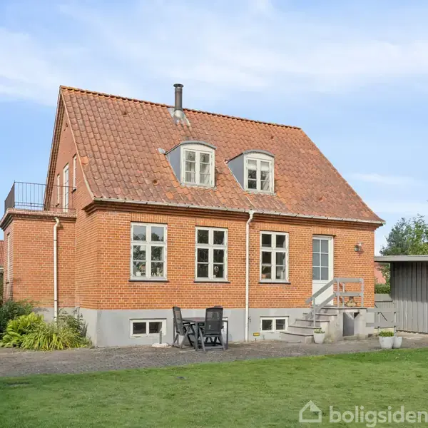 Rødt murstenshus med rødt tegltag; en havestol og et bord står foran huset. Græsplæne og buske omgiver huset. Der er en grussti og en lille terrasse.