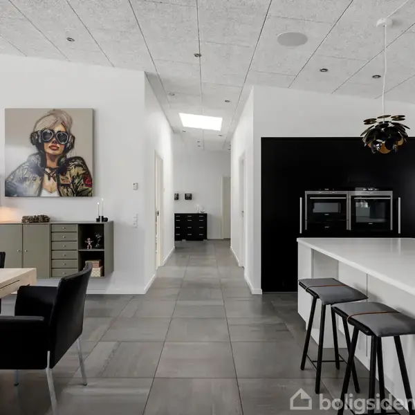 Køkkenområde med moderne indretning; en stor køkkenø med barstole til højre, og et langbord med stole til venstre; maleri på væg; lys og rummelig atmosfære.