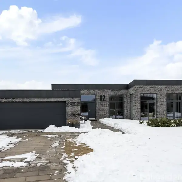 Et moderne hus med fladt tag står i et snedækket landskab. Husets facade er grå mursten, og nummeret 12 er synligt på væggen ved indgangen.