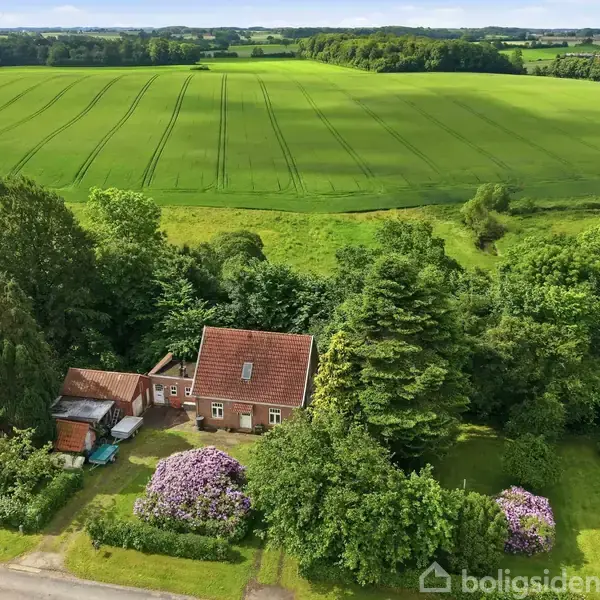 Et rødt hus med skråtag står omgivet af frodig grøn vegetation og store træer. Det ligger ved en vej med udsigt til store, åbne marker under en blå himmel.