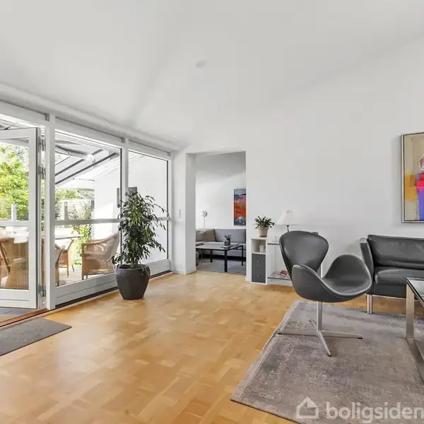 Stue med grå sofa og to moderne stole ved glasbord, står ved stor åbning til terrasse med stole. Vægge har farverigt maleri. Lyse trægulve.