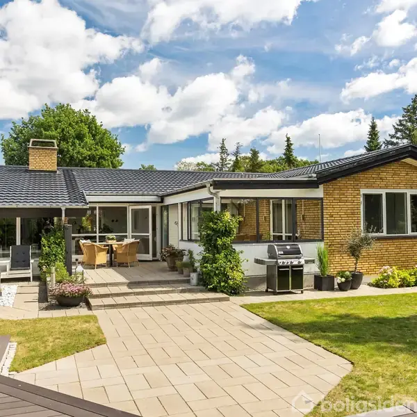 Et gult murstenshus med sort tag står i en velplejet have. Udendørs fliseterrasse med bord, stole og grill. Omgivet af græsplæne og buske under en blå himmel med skyer.