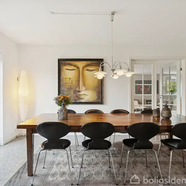 Et langt træbord står midt i en spisestue med seks sorte stole omkring det. En moderne lampe hænger over bordet, og en stor maleri hænger på væggen. Havedøre til venstre åbner ud mod en terrasse.