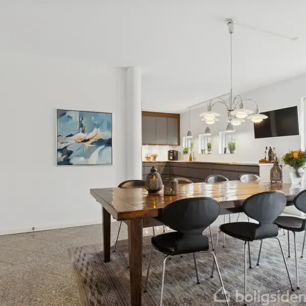 Spisebord omgivet af sorte stole står i et lyst køkken-alrum. Lamper hænger over bordet, og franske døre leder ud mod en grøn have. Maleri pryder væggen.