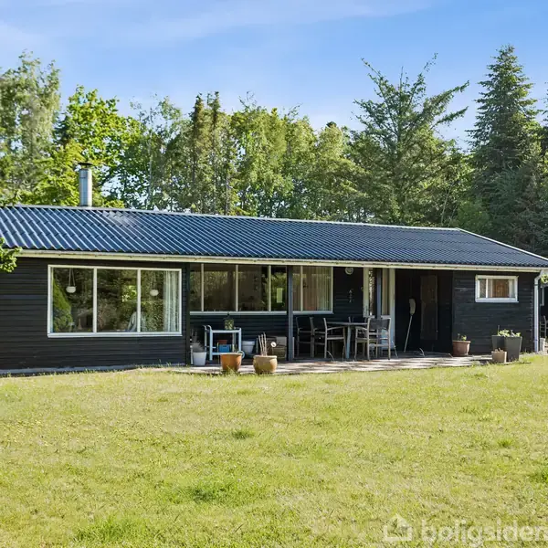 Et sort træhus med store vinduer står stille i en grøn have omgivet af træer. På terrassen foran huset står et bord og stole.