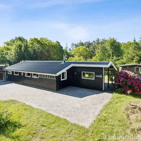 Et sortmalet hus står stille, omgivet af et grønt græsareal og tæt skov, med farverige blomster ved indgangen under en klar blå himmel.