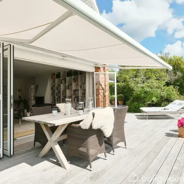 Udendørs træterrasse med spisebord og stole dækket af en stor markise, omgivet af have og grønne planter. Terrassegulvet er lyst og knyttet til hvidt murstenshus med åben skydedør.