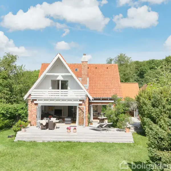 Et rødt hus med spids tag står stille i en grøn have omgivet af træer under en blå himmel med skyer. Der er en udendørs terrasse foran.